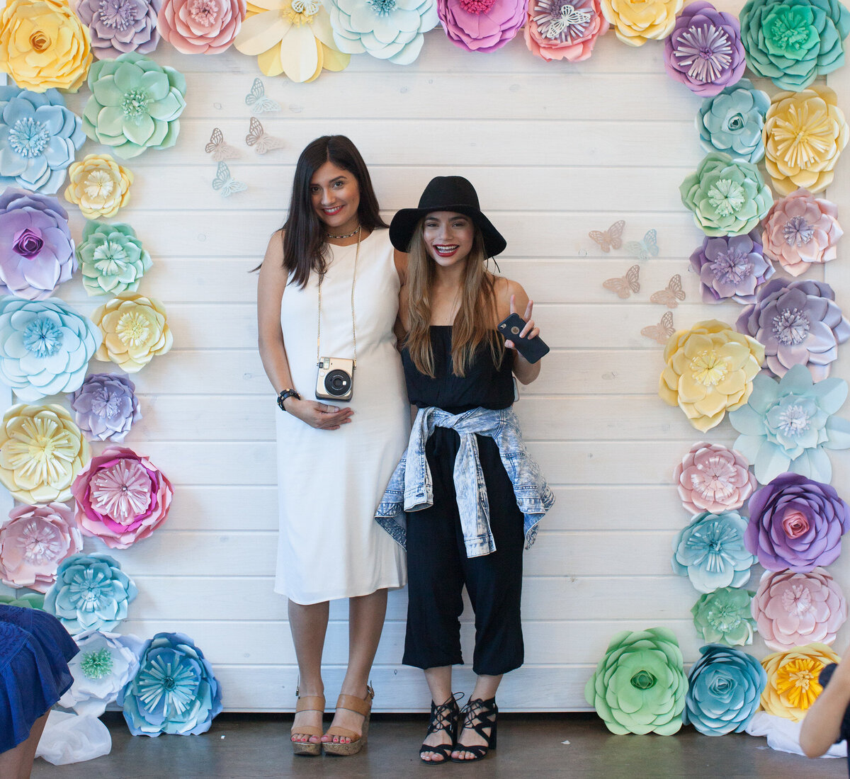Two women posing in front of a white backdrop decorated with large colorful flowers and butterflies