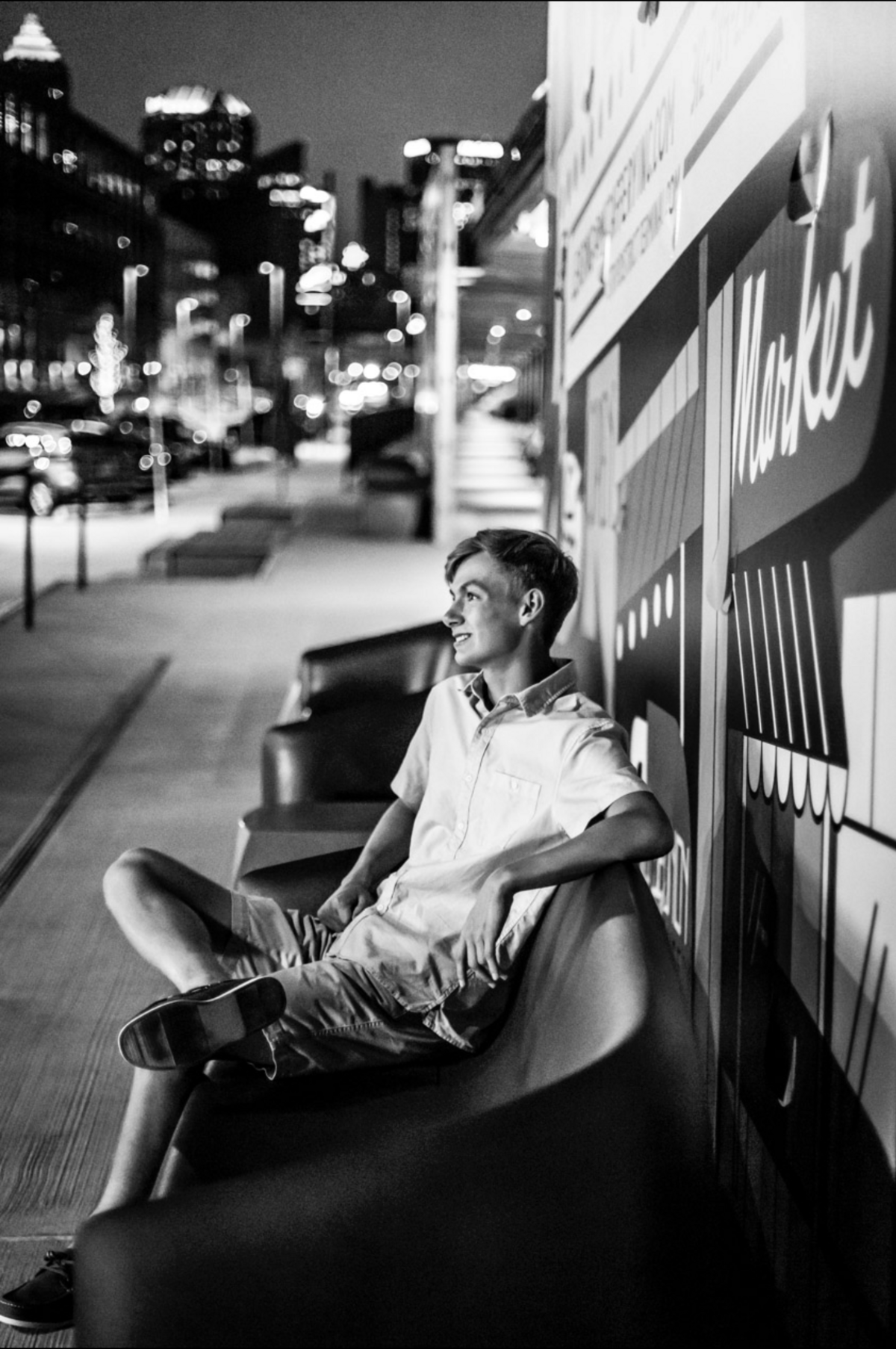 A black-and-white night time image of a senior boy sitting on a bench with the city Pittsburgh skyline in the background.