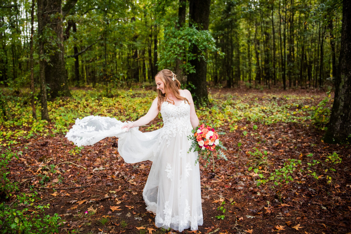 A bride fluffs the train of her wedding dress, standing in a woodland setting.\