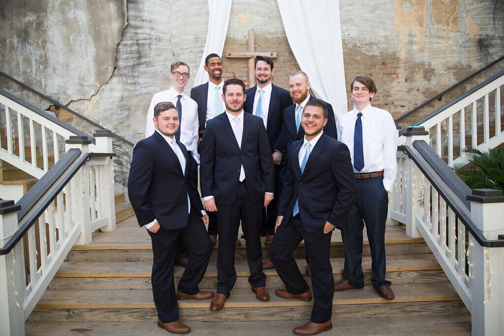 A group of eight men in dress shirts, ties, and suits pose together on outdoor wooden stairs with white railings, in front of a brick wall draped with white fabric and a wooden cross.