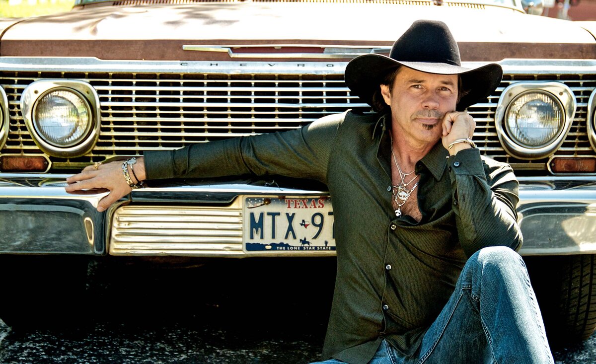 Country musician portrait Derryl Perry wearing black cowboy hat with green shirt sitting against old car front