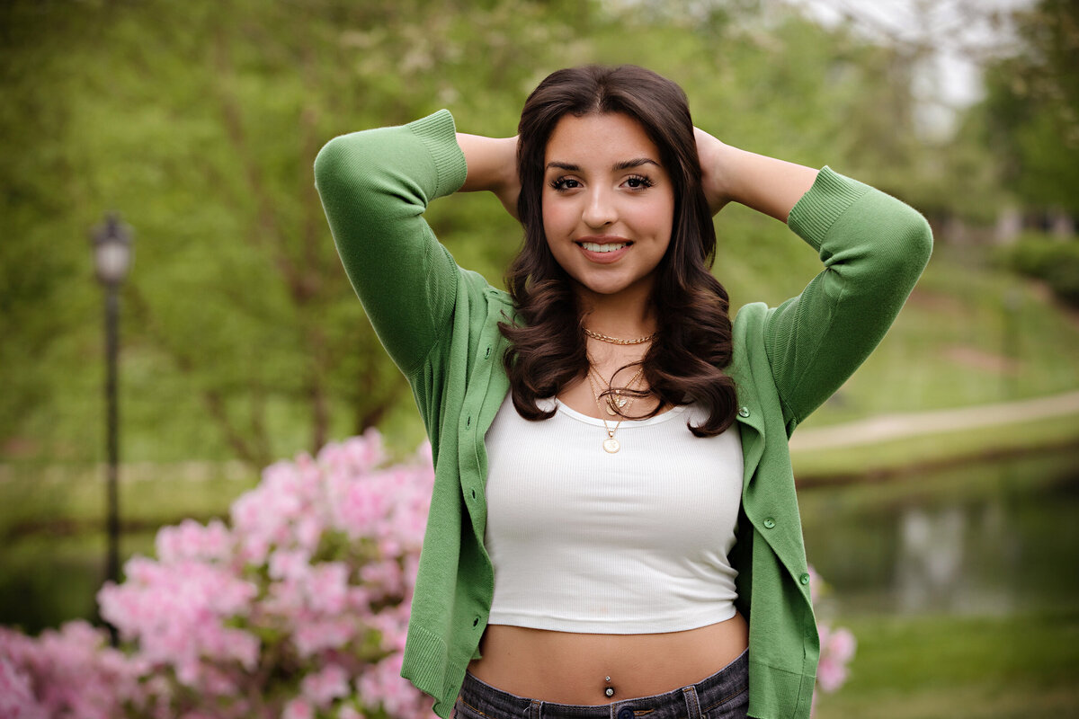 Standing senior portrait with arms raised holding hair, wearing a green cardigan and gray jeans in a spring garden setting.