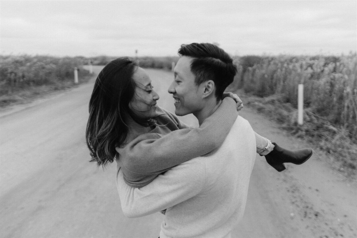 The man is carrying his fiancee on the road, amidst tall, dry grasses in Fire Island Beach, NY. Engagement Image by Jenny Fu Studio