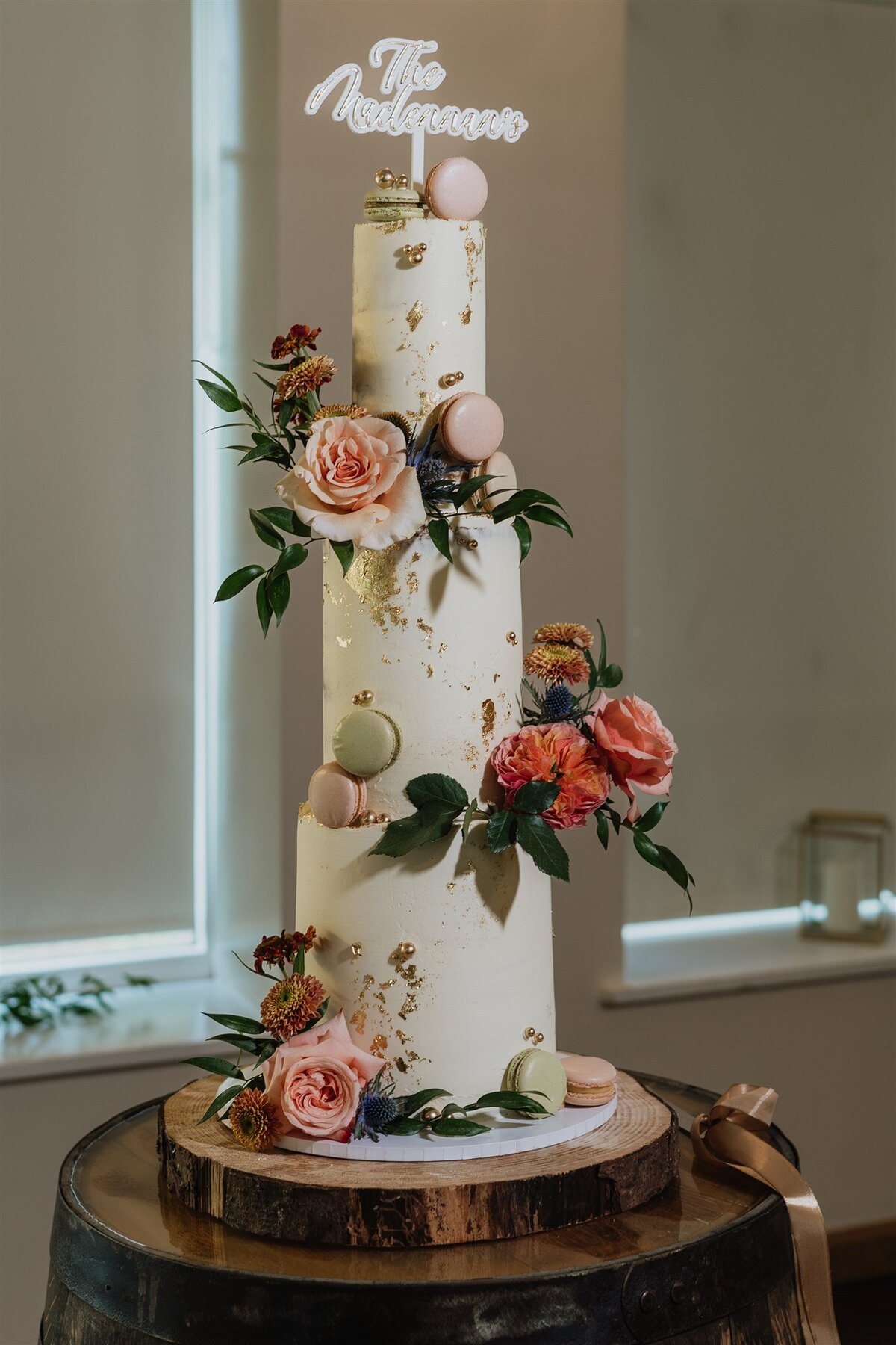 A three tier wedding cake stands on a rustic barrel. The cake is decorated with cream icing, roses, macarons, and foliage.