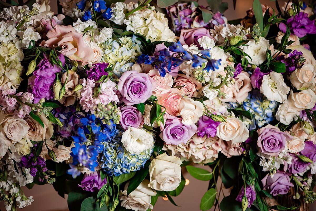 A large floral arrangement with light purple roses, dark purple flowers, dark blue flowers, white roses, pink roses, blush roses, blue hydrangea, white hydrangea and greenery.