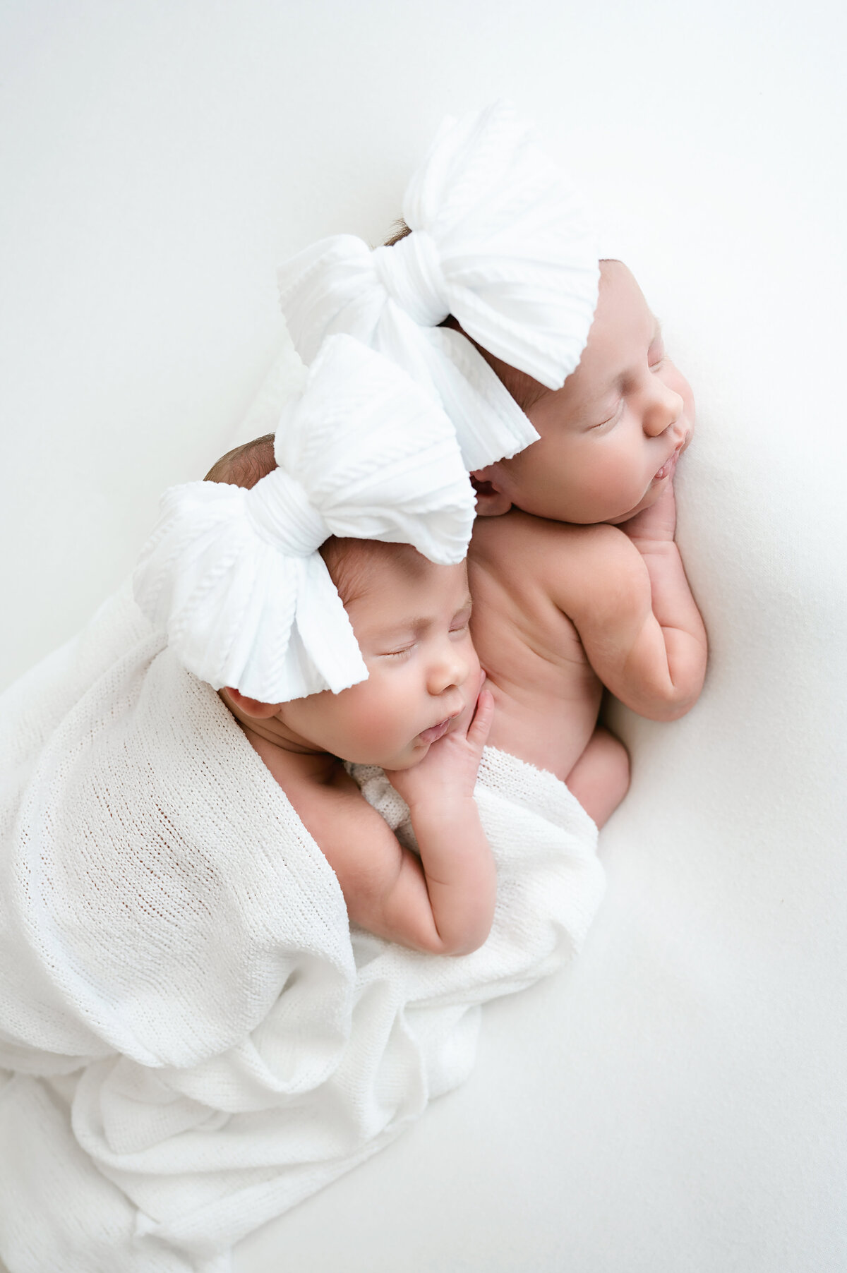 A precious twin newborn photography session in Pittsburgh, capturing two sweet babies bundled in soft white blankets with matching large white bows. This serene image celebrates the beauty and tranquility of newborn twins during their first days