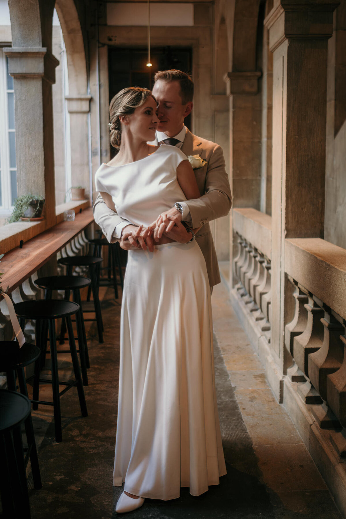 The groom hugs his bride from behind her, while standing in a historical venue.