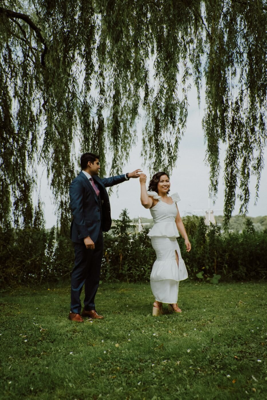 A full-body candid shot of a couple standing in a park under a large weeping willow tree. The man, in a blue suit with a pink tie, gently holds the woman's hand, who is playfully spinning or dancing. The woman wears a white two-piece outfit with ruffles and a slit. The ground is covered in lush green grass and clover, with a body of water visible in the background.