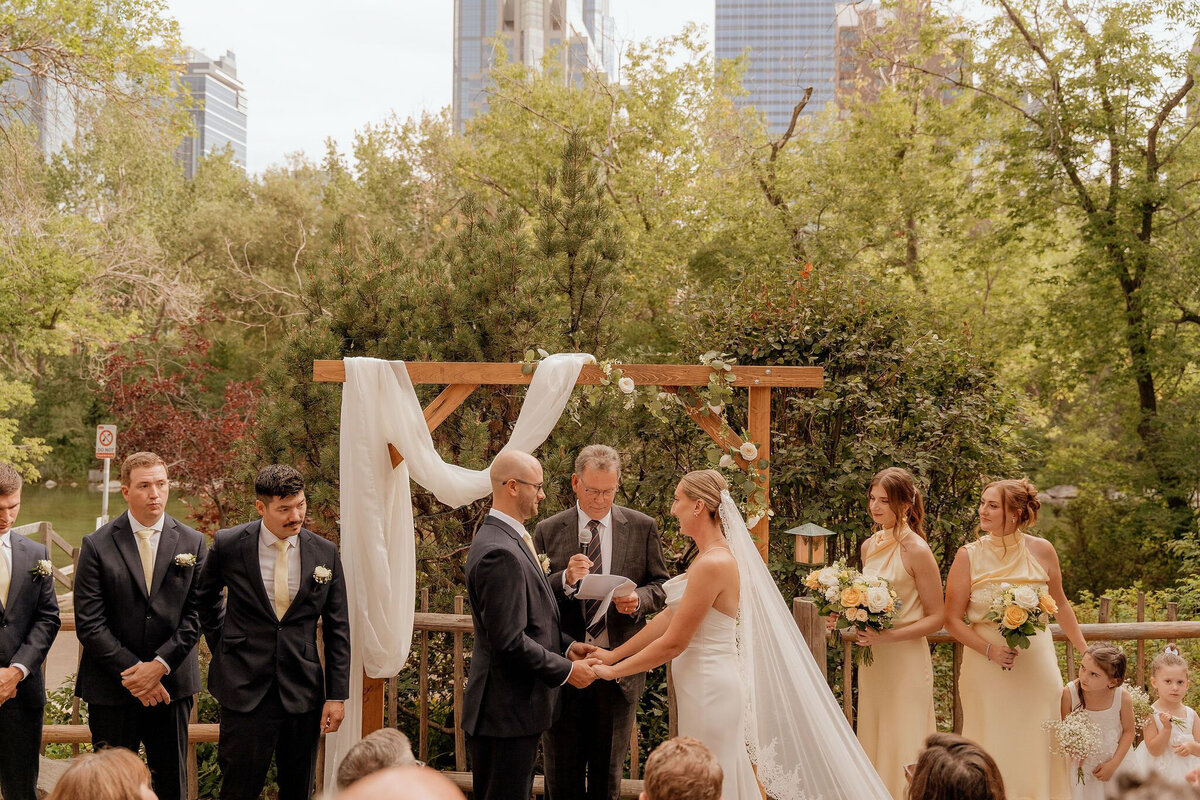 Outdoor wedding ceremony at River Café in Calgary, Alberta, featuring a couple exchanging vows under a vibrant floral arch. Surrounded by lush greenery, the bride in a flowing white gown and the groom in a classic black suit stand before their officiant. Bridesmaids in deep green dresses and groomsmen in black suits add to the elegant scene, while guests watch from floral-adorned wooden chairs. A romantic and picturesque setting along the Bow River.