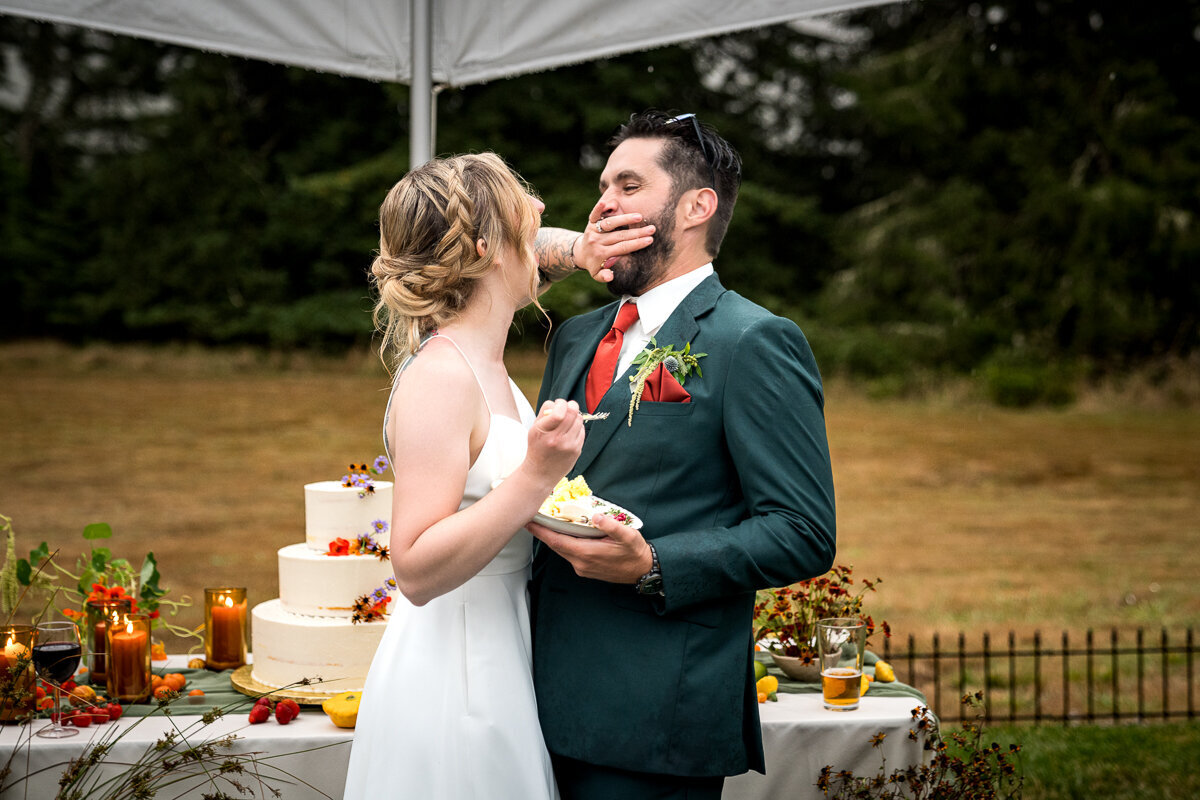 A lighthearted and fun moment as the bride playfully shoves cake into the groom's mouth during their reception at Ridgefield on Fickle Hill in Arcata, surrounded by laughter and joy.