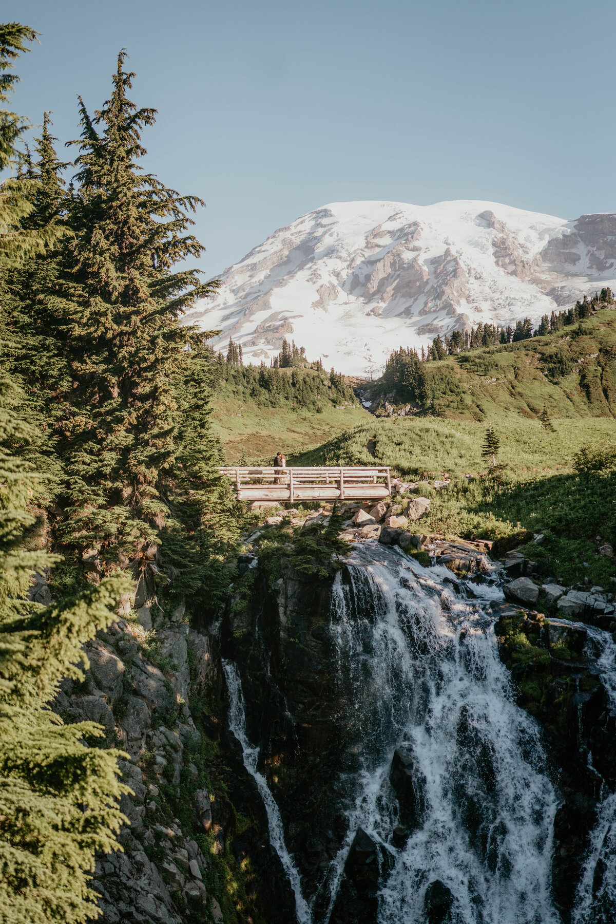 Tiny couple crossing a bridge at Myrtle Falls with views of Mt Rainier