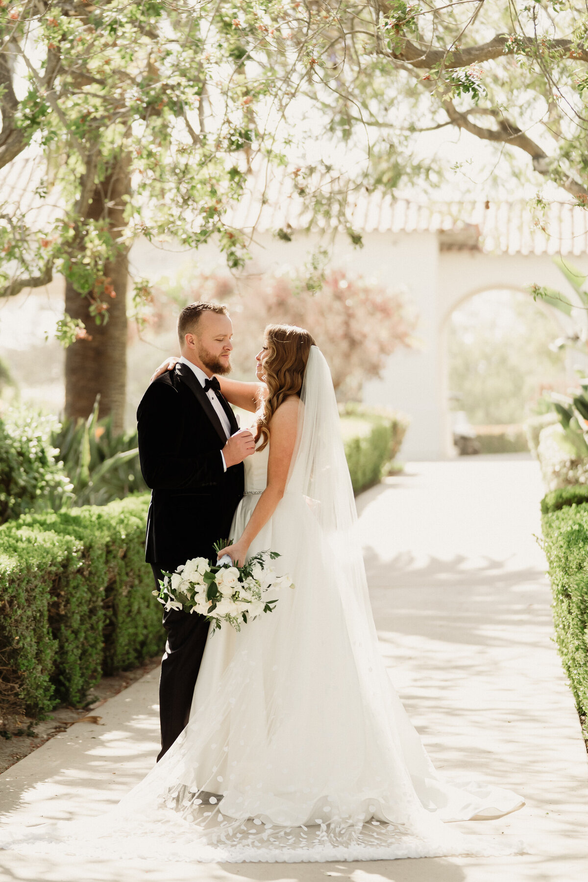 groom in a black tux with a bowtie and bride in a white dress with a cathedral polka dot veil with a large white bouquet talking during romantic wedding photography at spanish hills country club captured by los angeles wedding photographer magnolia west photography