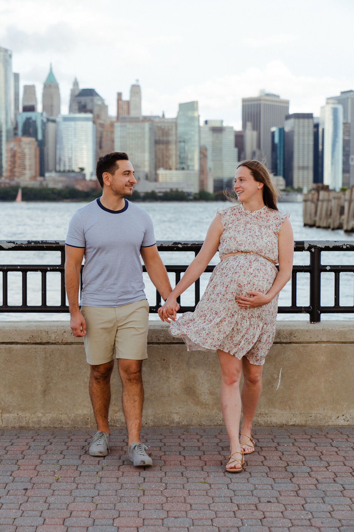 a couple walks together in jersey city with the skyline view of manhattan behind them