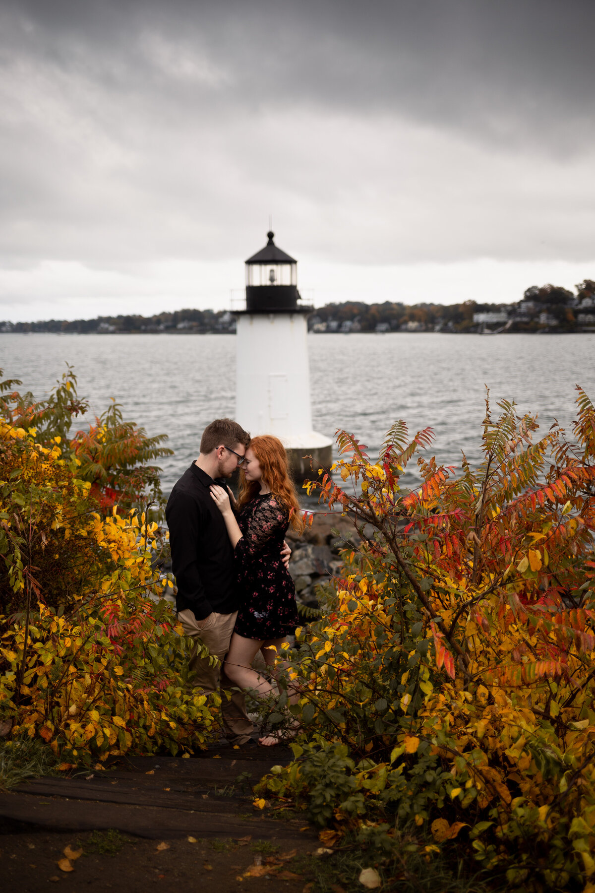 A couple cuddles one another in front of a lighthouse surrounded by the ocean.
