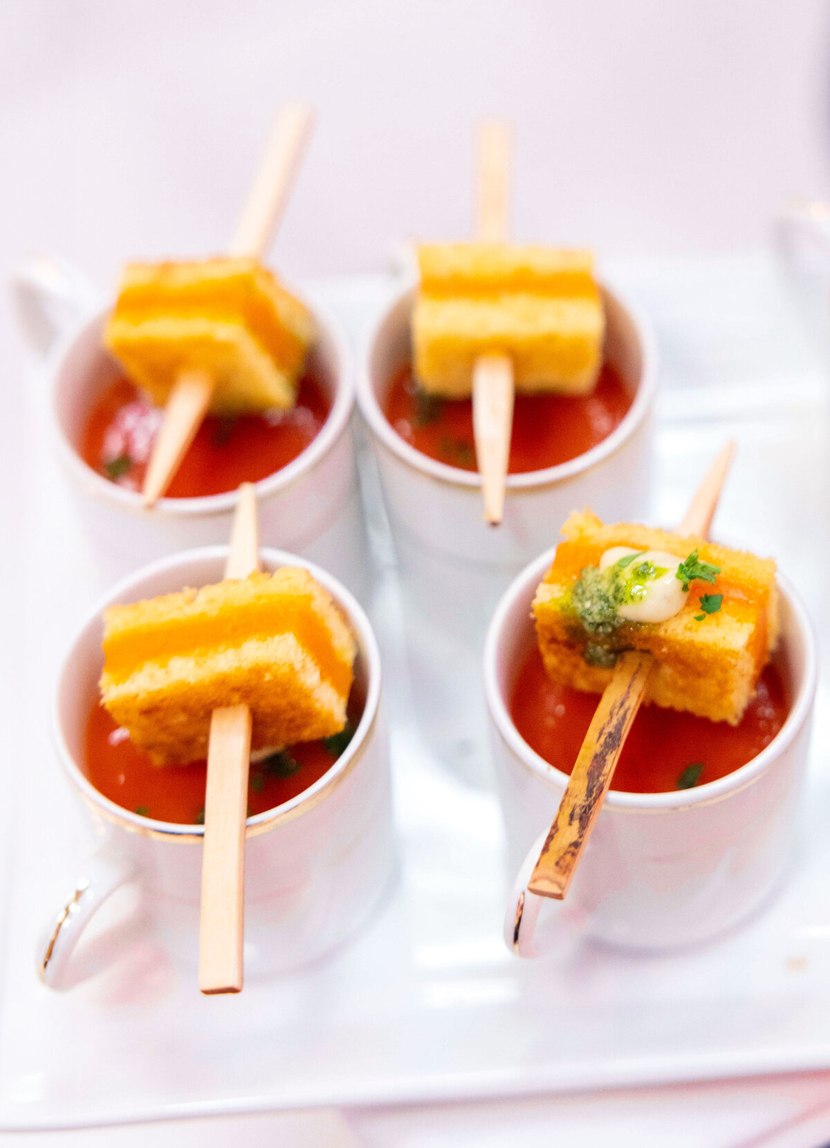 Four white cups filled with tomato soup, each garnished with a skewer of grilled cheese sandwiches, sit on a white platter. The skewers are topped with a dollop of cream and a sprinkle of herbs.