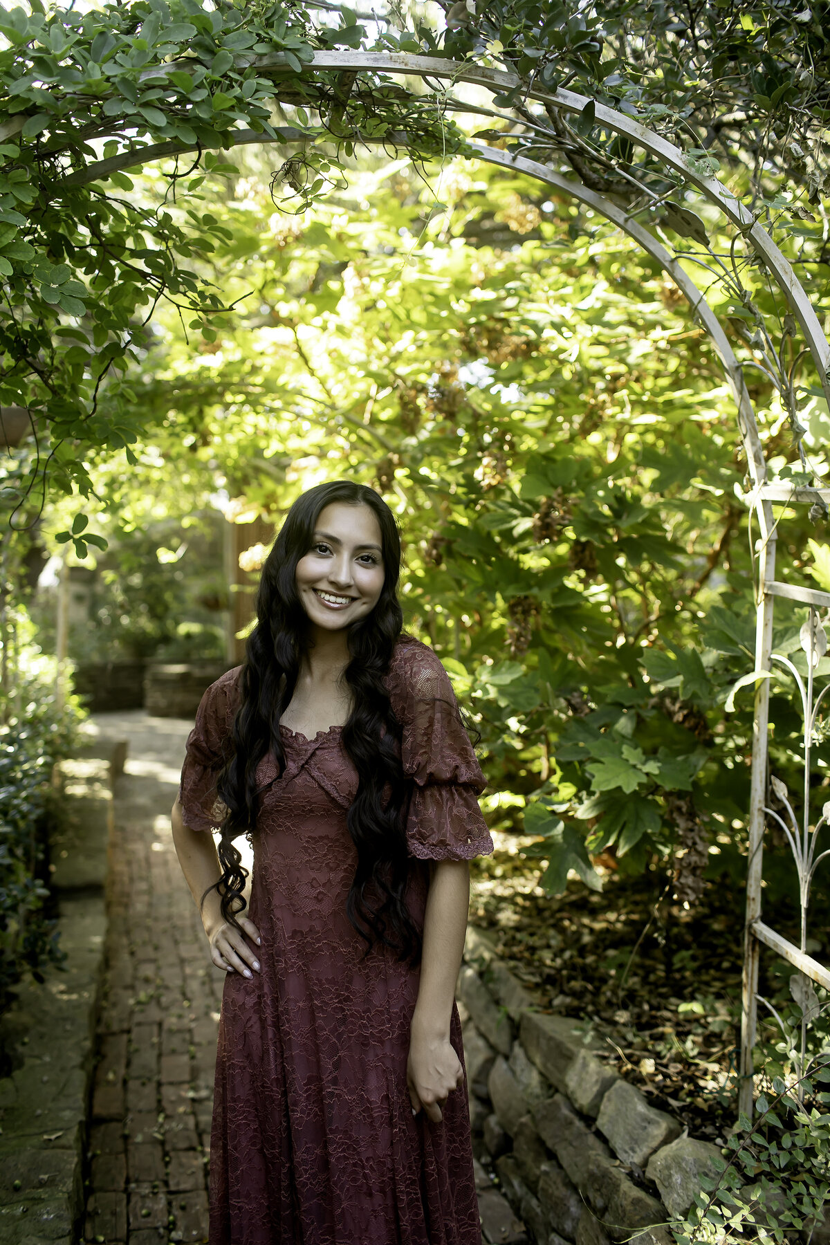 A high school senior smiling brightly in the breathtaking landscape of Chandor Gardens in Weatherford, TX, with the blooming flowers adding a pop of color to the moment