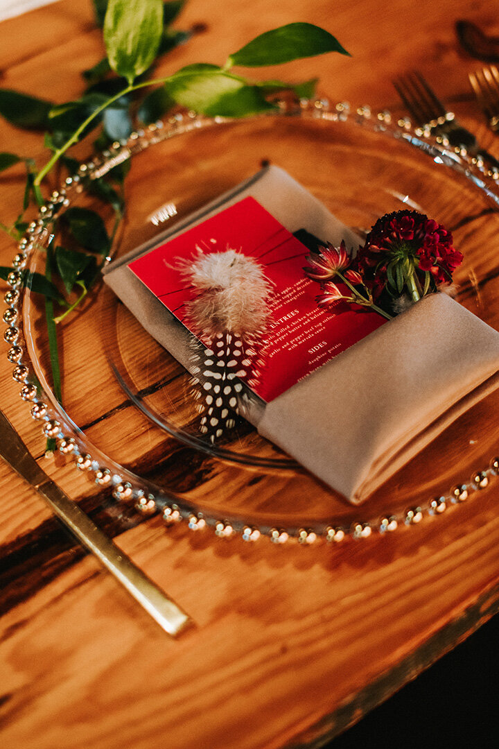 Red card stock dinner menu set atop a gold napkin with flowers on a clear plate and gold silverware.