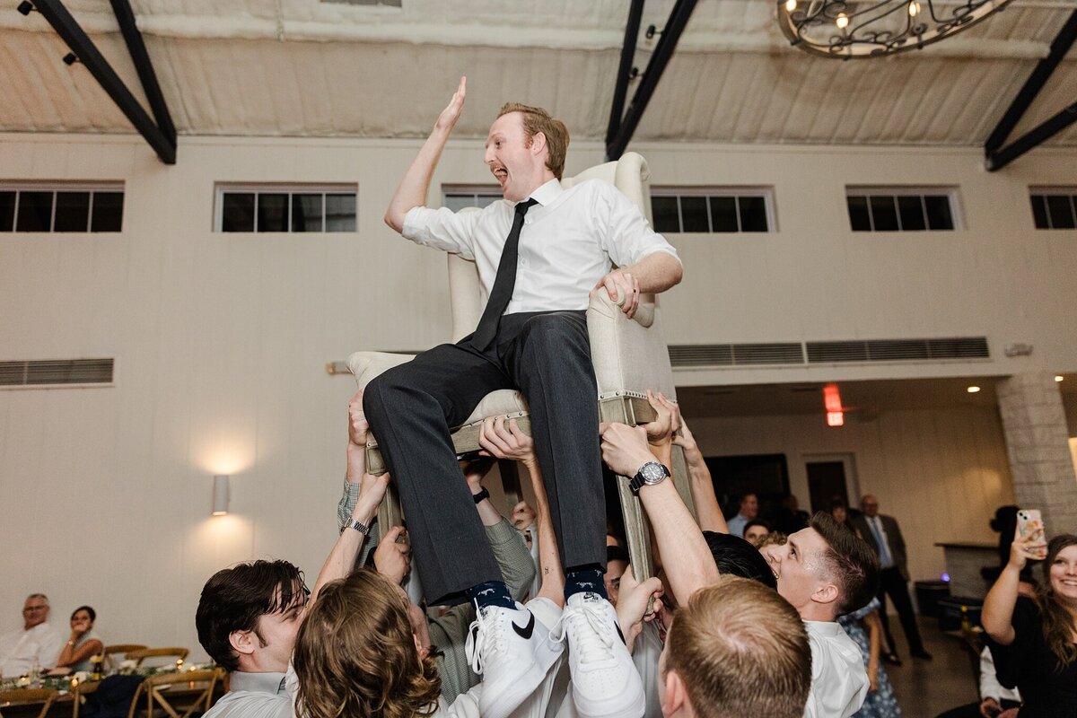 A groom being held up in the air by wedding guests while sitting in a chair during his wedding reception at Bella Cavalli Events in Aubrey, Texas. The groom is wearing a suit minus the jacket and is joyously cheering as he's hoisted around the reception hall.