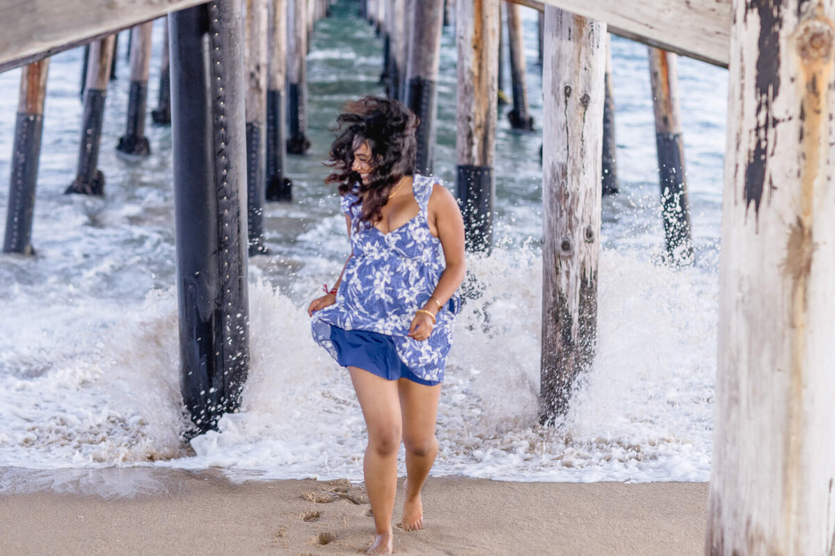 A person in a blue and white dress runs barefoot along a sandy beach, under a wooden pier with crashing waves in the background.