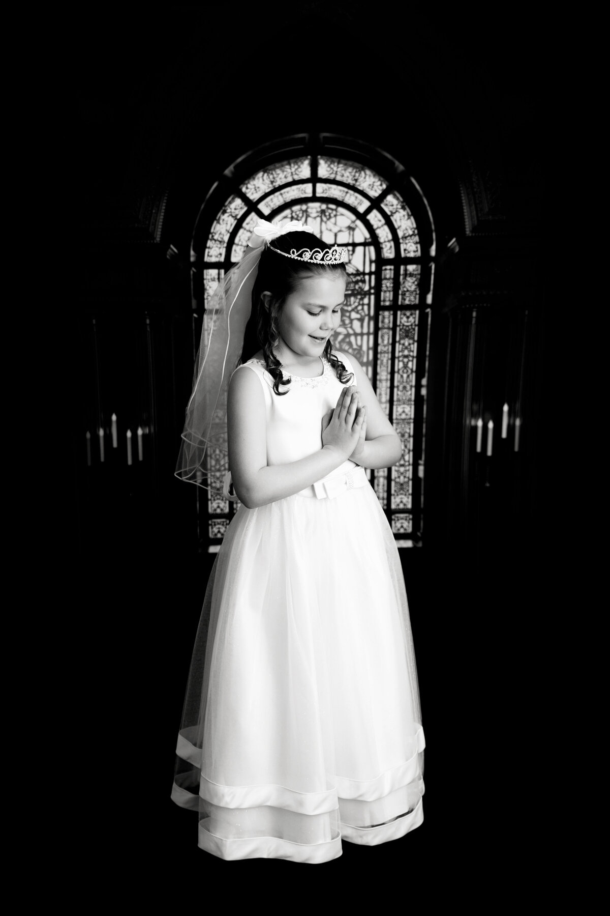 A black and white photo from a New Jersey Communion Portrait Photographer of a young girl in a dress and tiara praying in front of stained glass window