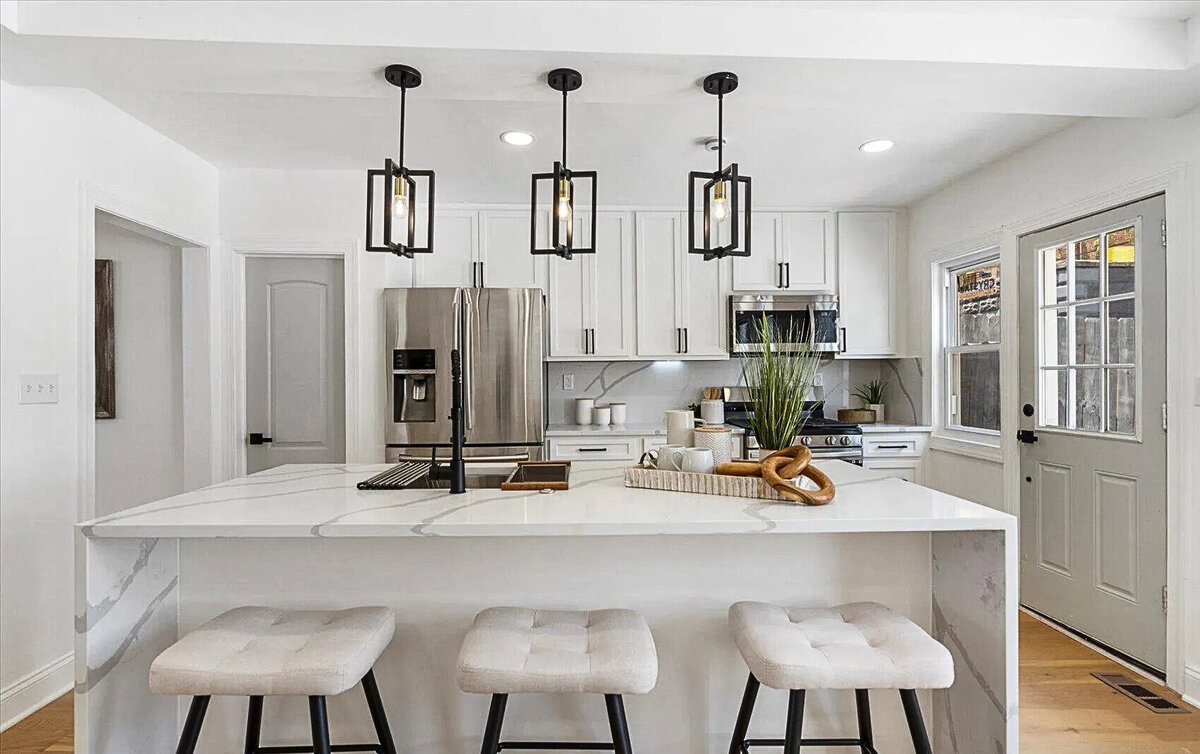 Modern white kitchen with marble island, black pendant lights, stainless steel appliances, and bar stools.