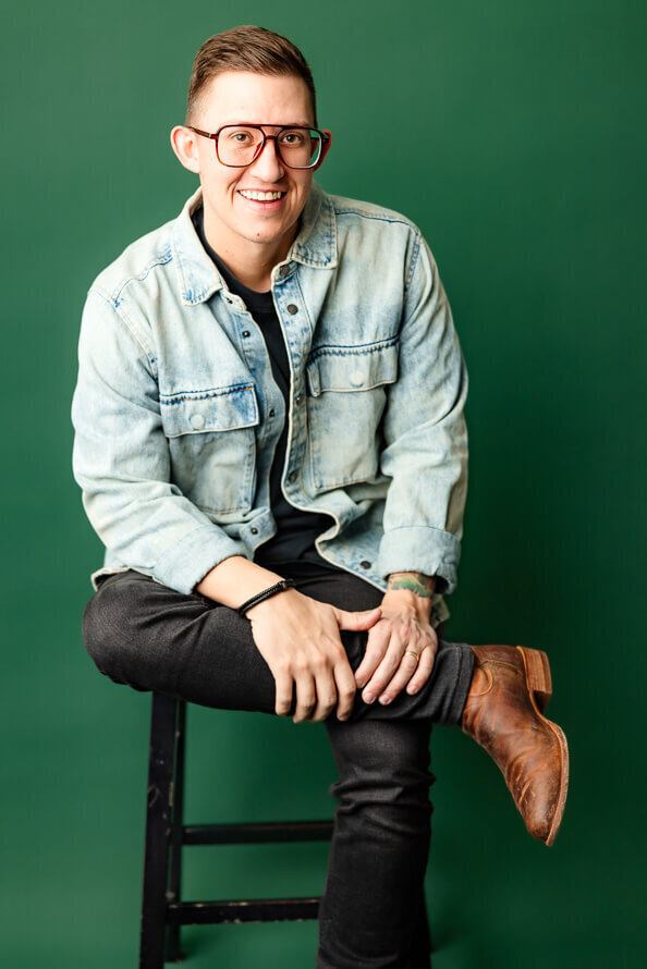 Man in jean jacket sits and holds onto leg for his headshot session.