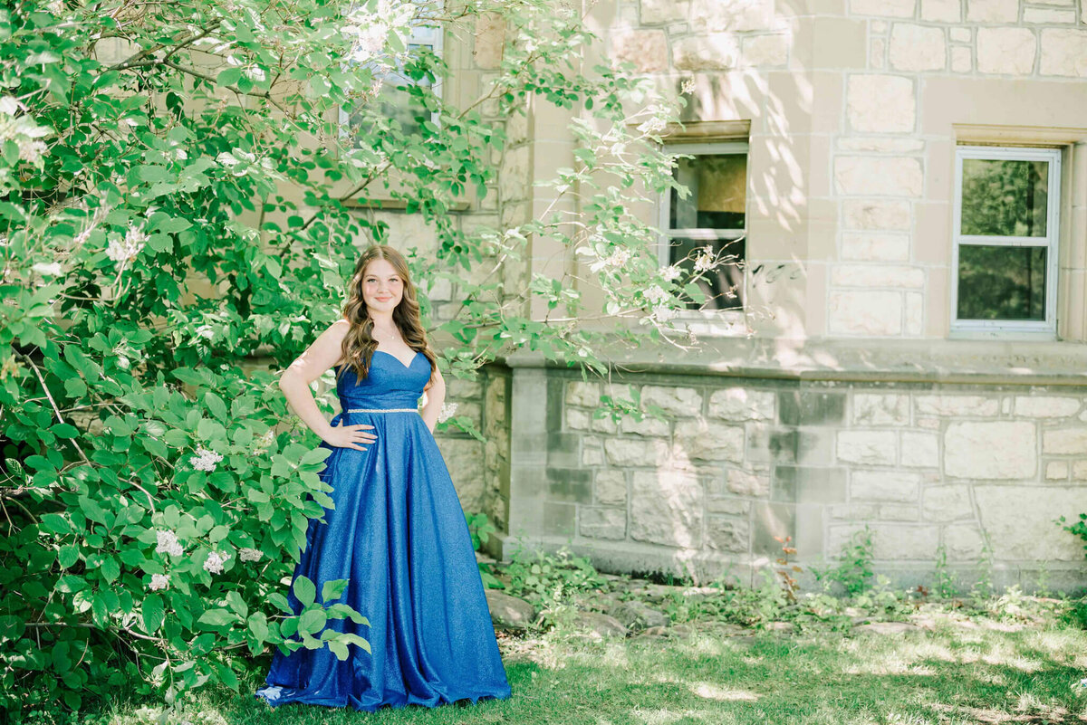 Standing with her back to trees and beside and building. 