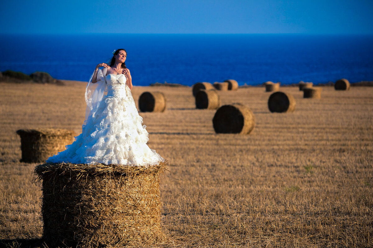 Bride standing on a hay bale against a backdrop of a feild of hale bales with the sea in the distance