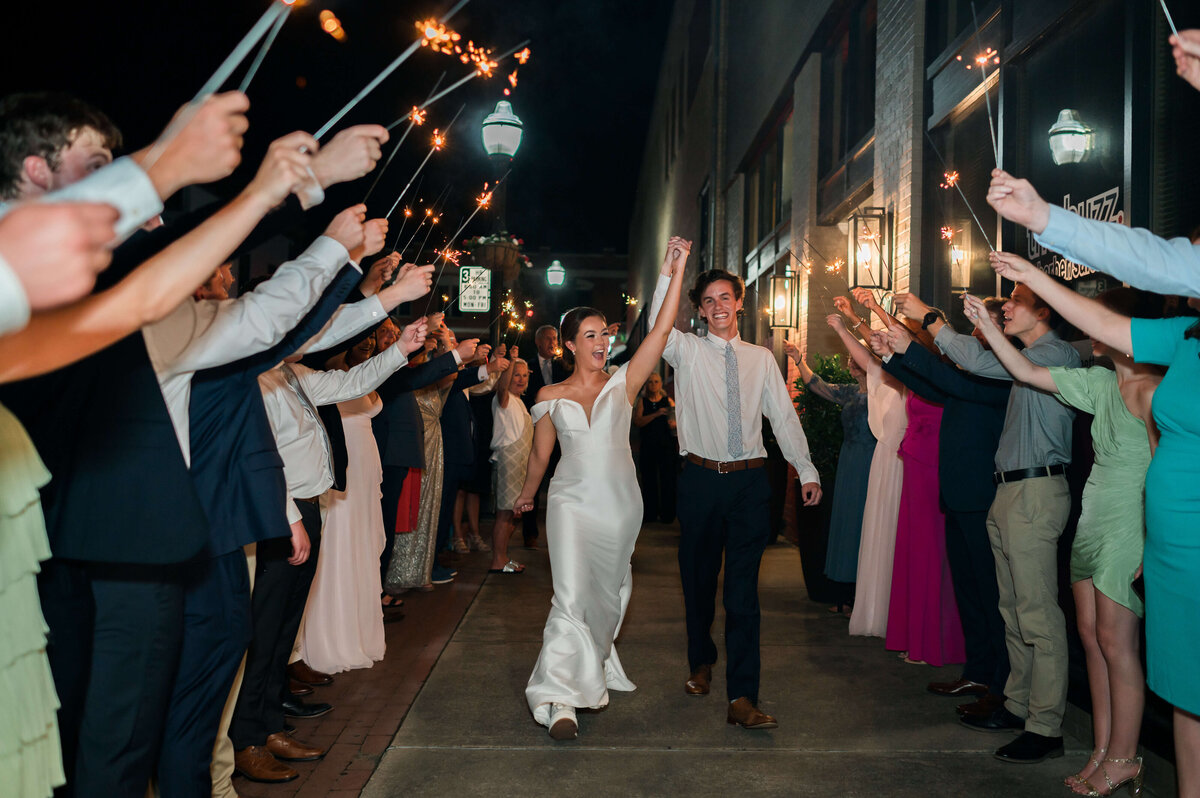 A bride and groom smile and hold hands while walking through a cheering crowd holding sparklers at night, celebrating their wedding on a city sidewalk.