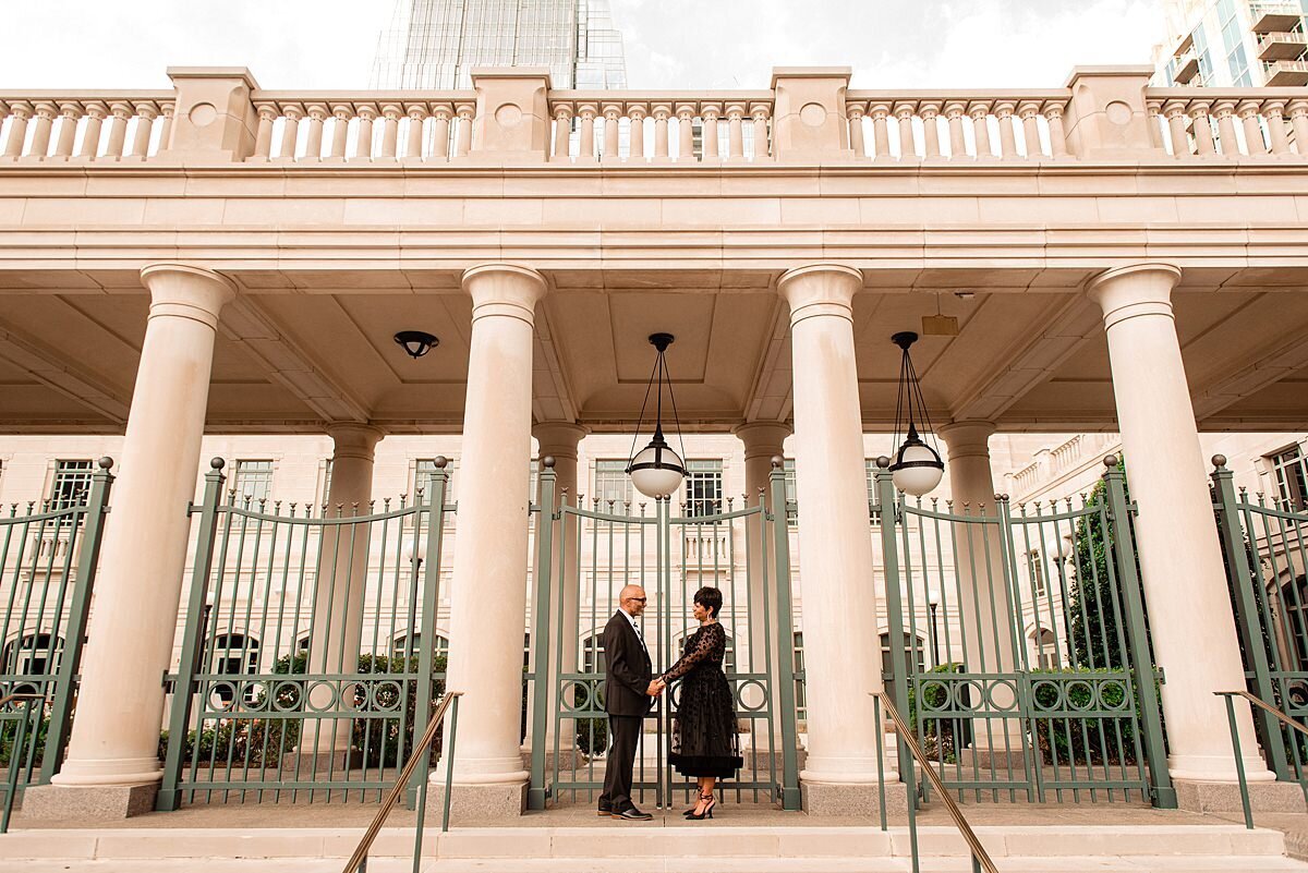 The bride and groom stand in a colonnade  with tall metal gates behind them the bride and groom hold hands as they look at each other. The groom is wearing a black tuxedo and the bride is wearing an elegant black tea length lace dress.