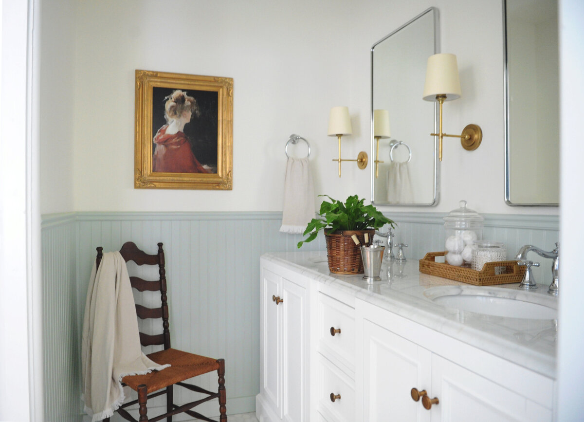 Traditional bathroom design with blue wainscoting and white vanity. Bathroom has wooden accent chair with gold framed portrait and brass sconces and mirror.