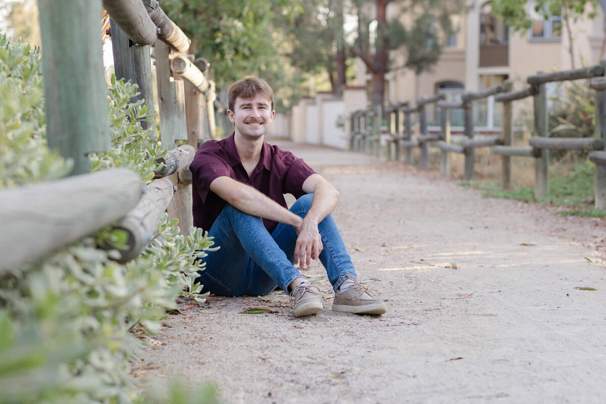 A young man with dirty blonde hair and a mustache leaning against a wooden park fence, sitting on the gravel ground of a park walking path, draping his arms over his knees and smiling at the camera from afar.