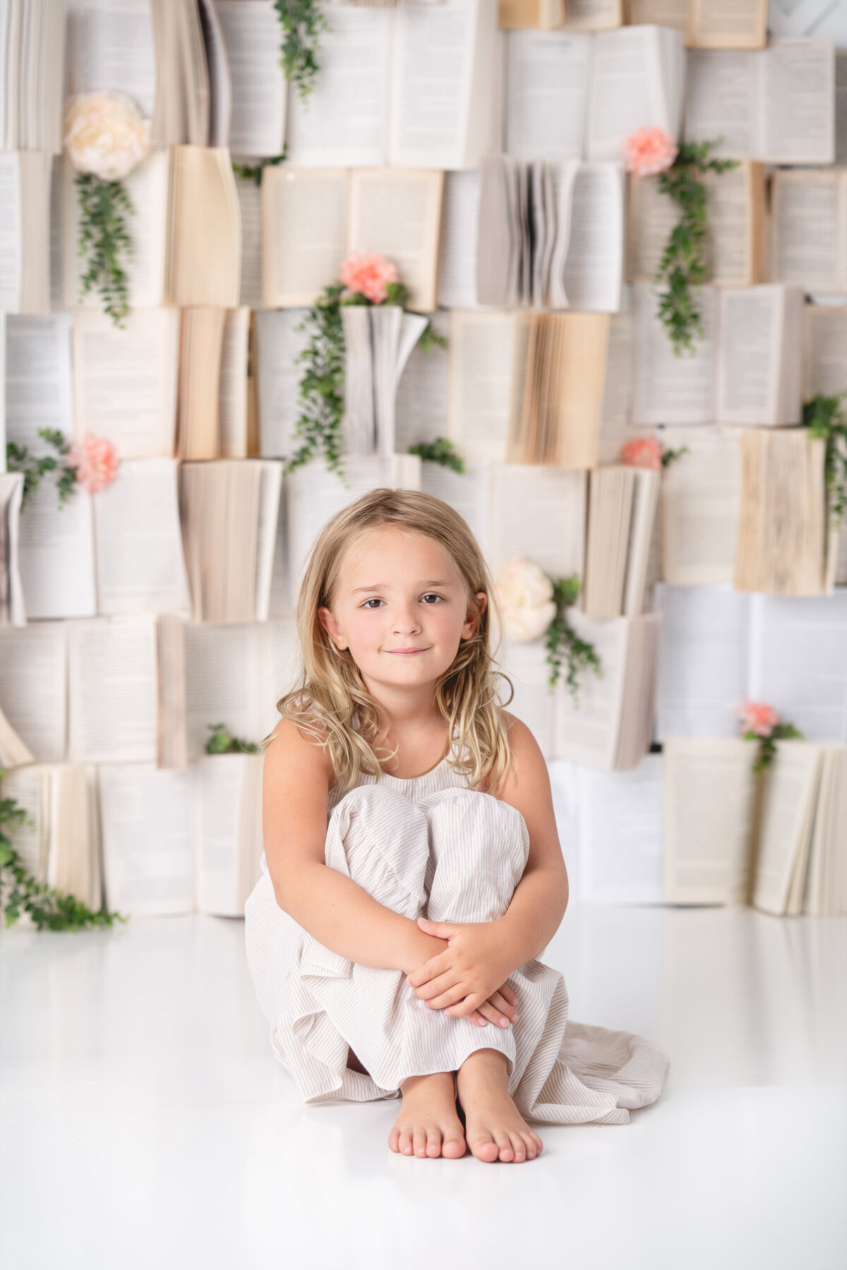 A young girl is sitting in front of a backdrop made of books.
