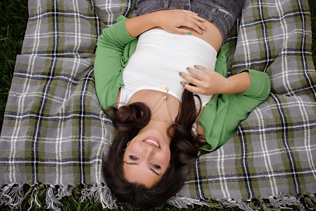Senior portrait of teen lounging on a blanket in the grass with soft spring light and layered textures.