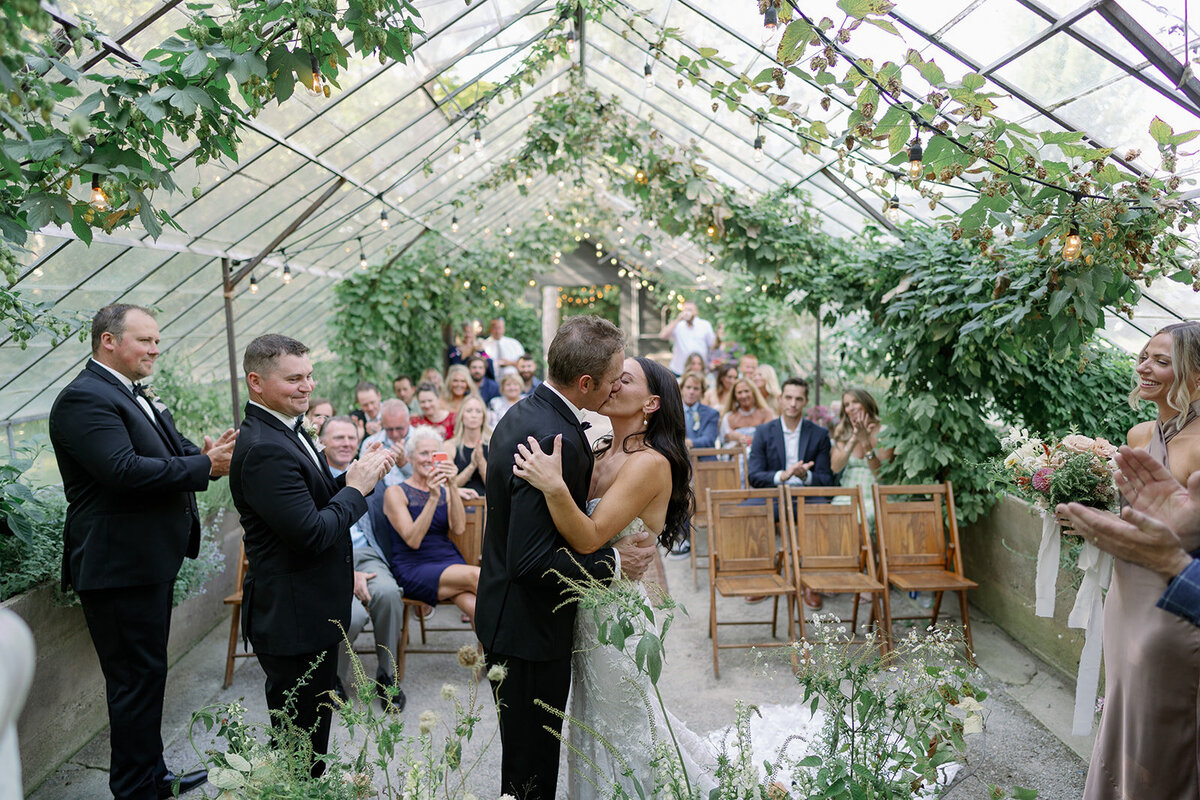 Bride and groom kissing during their ceremony inside the greenhouse at Glasshouse Community, a popular Michigan wedding venue.