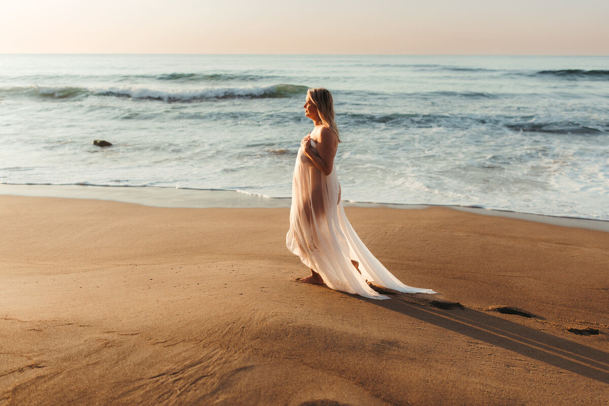 Pregnant woman standing barefoot on the shoreline at sunset, draped in a sheer flowing gown, with soft golden light casting long shadows on the sand and waves gently rolling in behind her.