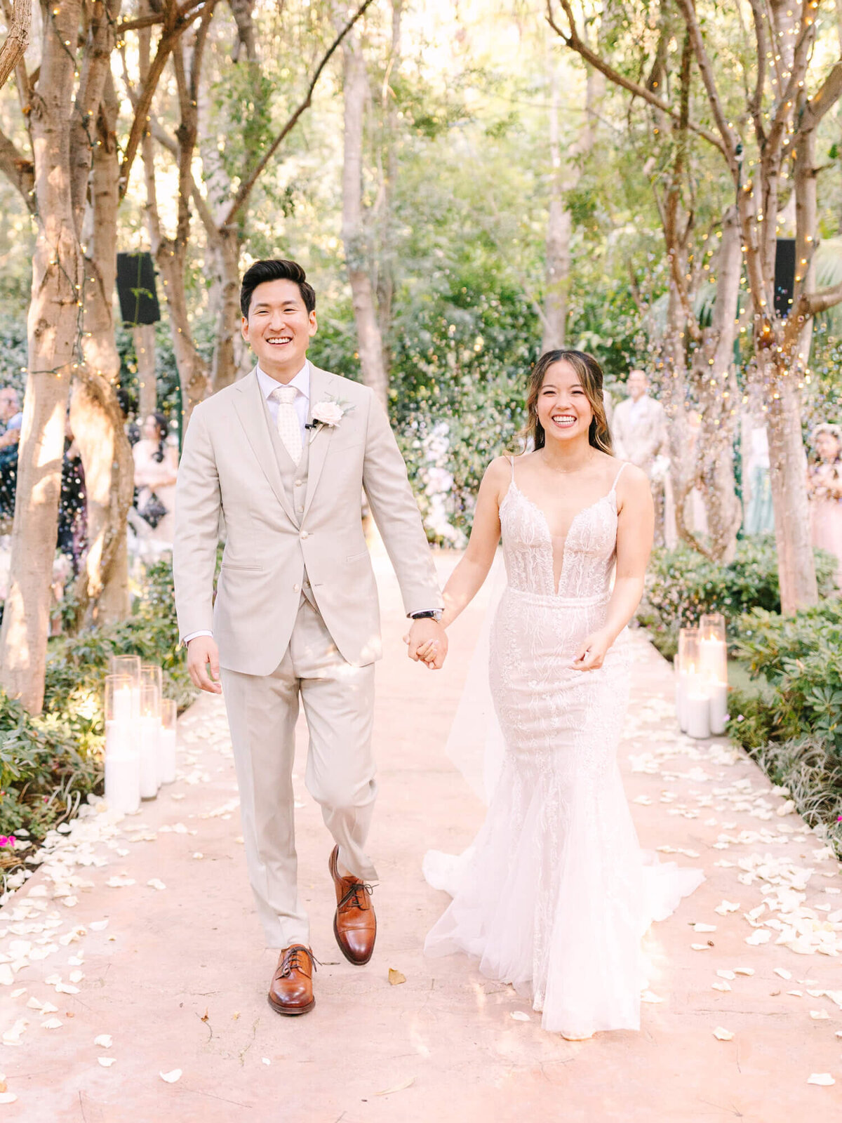 A joyful couple walks down an outdoor aisle surrounded by trees and rose petals.