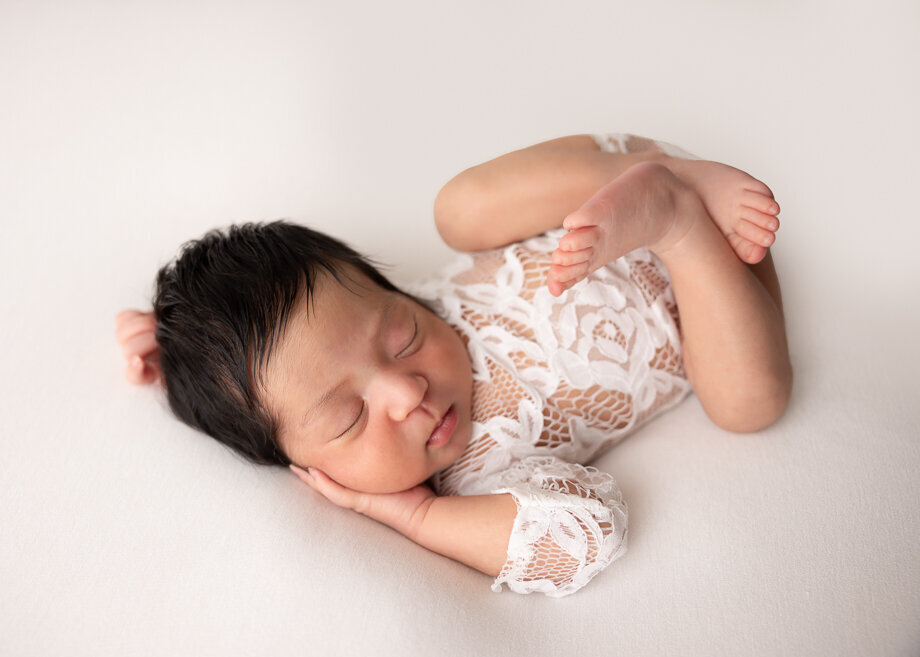 This baby is posed on her back in a lace bodysuit on a clean white backdrop.  She has a  hand tucked under her chubby cheek and legs are tucked up in a huck fin pose.