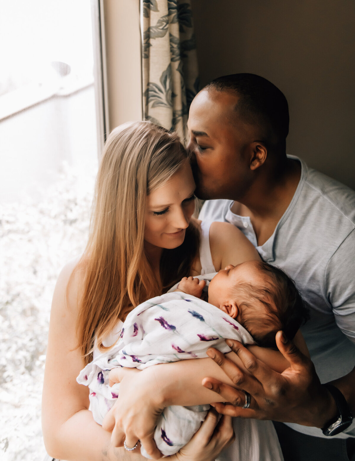 Newborn Photographer, a mother holds her newborn baby beside the window, dad kisses mom