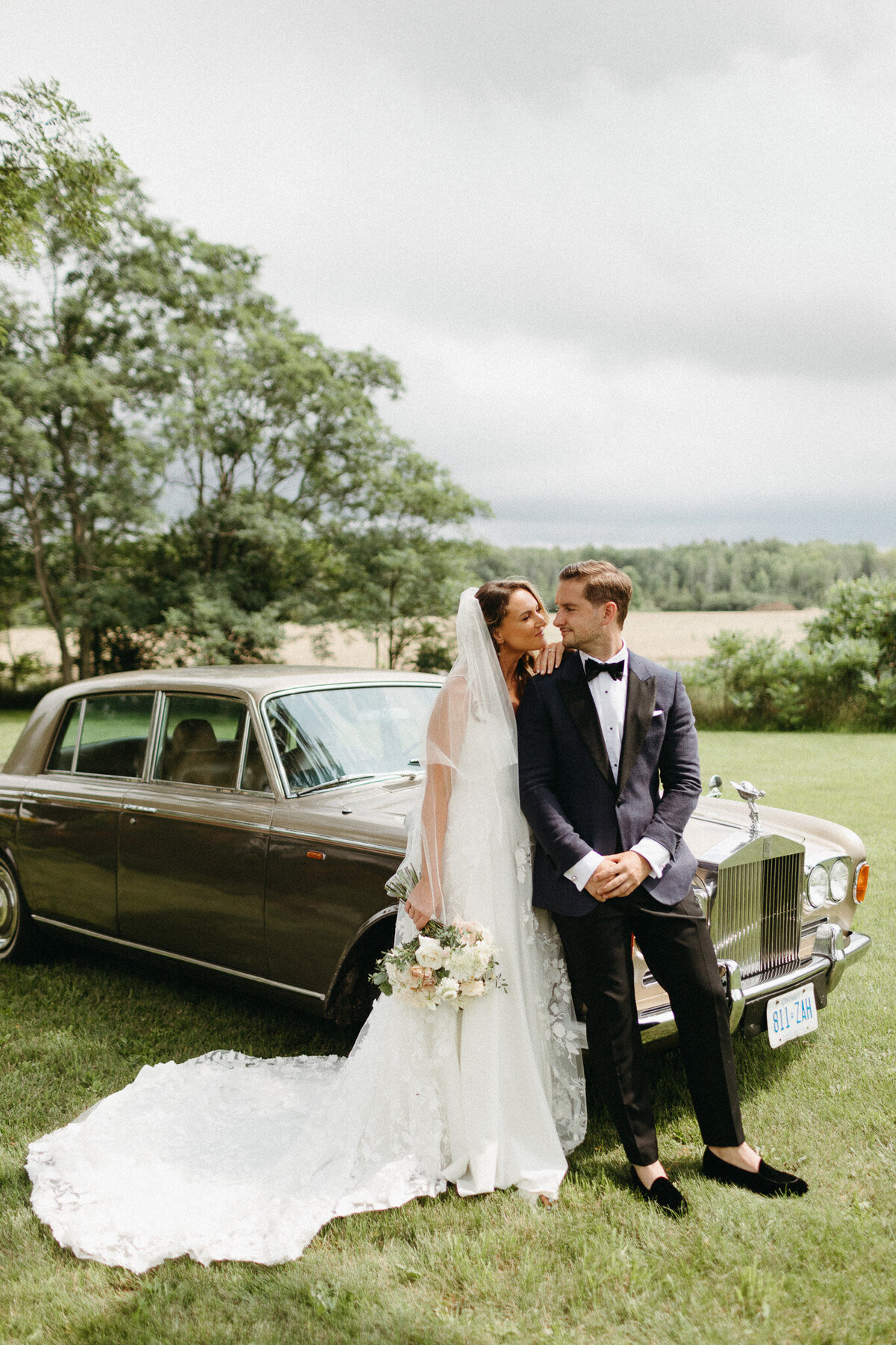 Bride in a white gown and groom in a tuxedo stand by a vintage car in a grassy field, captured beautifully by a talented Muskoka wedding photographer, with trees and cloudy sky enhancing the romantic backdrop.