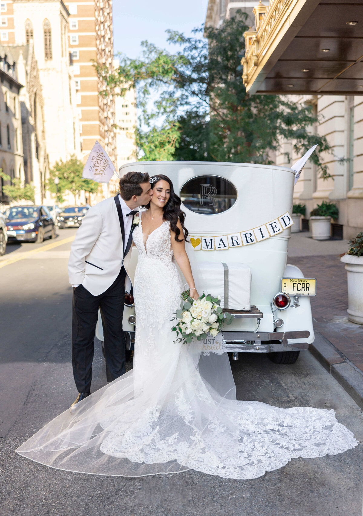 Bride in a lace gown and groom in a white tuxedo stand beside a vintage car with a Just Married sign. The groom kisses the brides cheek. They are on a city street, surrounded by buildings and trees. The bride holds a bouquet of white flowers.