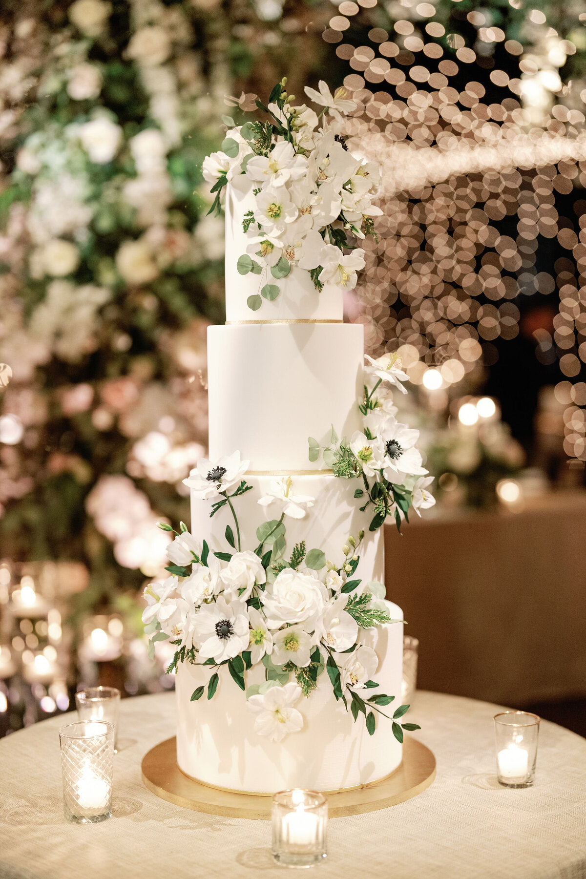 A stunning multi-tiered white wedding cake adorned with delicate sugar flowers, set against a backdrop of twinkling lights and lush greenery.