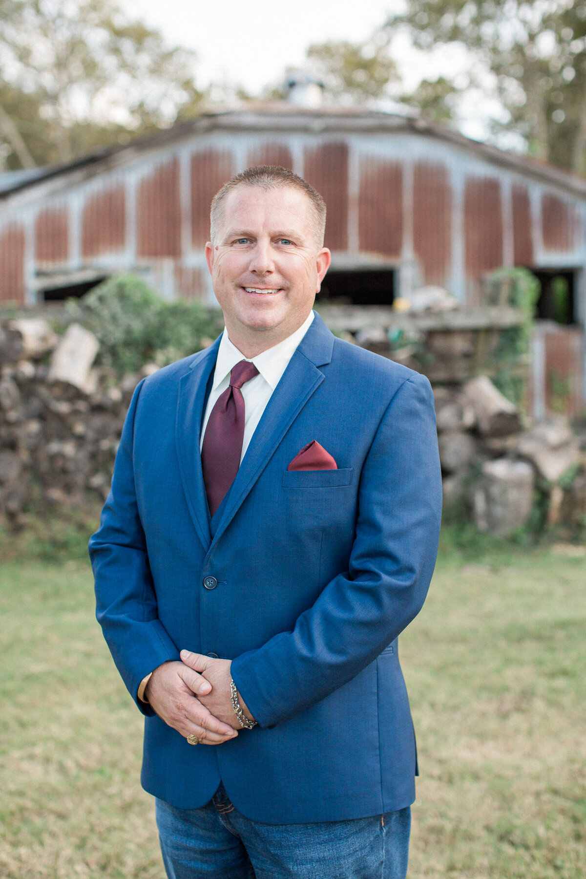 The groom stands with folded hands in front of a wood pile and rustic barn