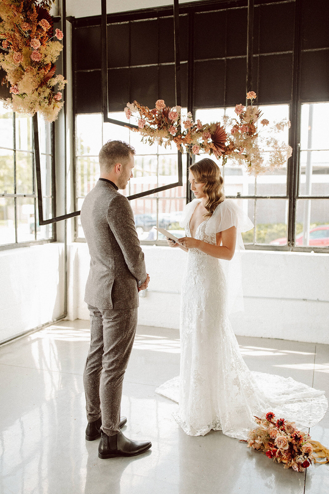 Bride and groom wearing a gray suit and white wedding gown stand facing each other in light airy room filled with flowers.