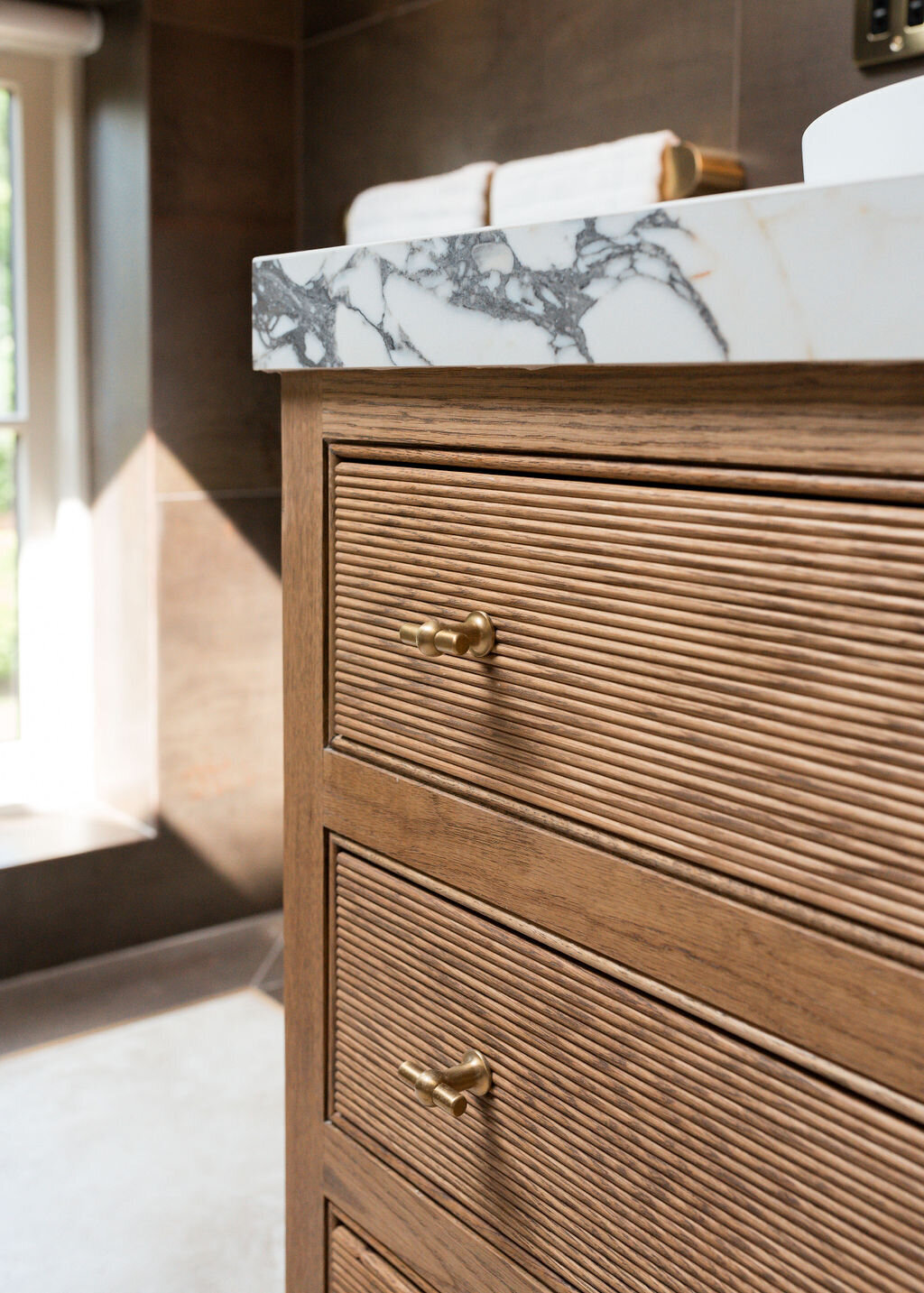 A close-up view of a wooden bathroom vanity with ribbed drawer fronts and brass drawer pulls. The countertop features white and gray marble. A bright window illuminates the space in the background.