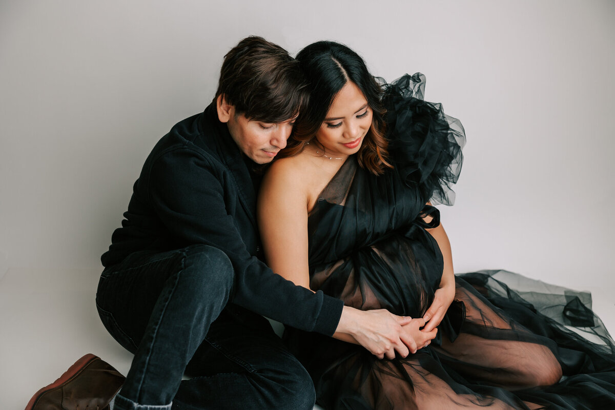 maternity portrait of a couple sitting down with their hands on her belly. She is wearing a black dress and he is wearing black sweater.