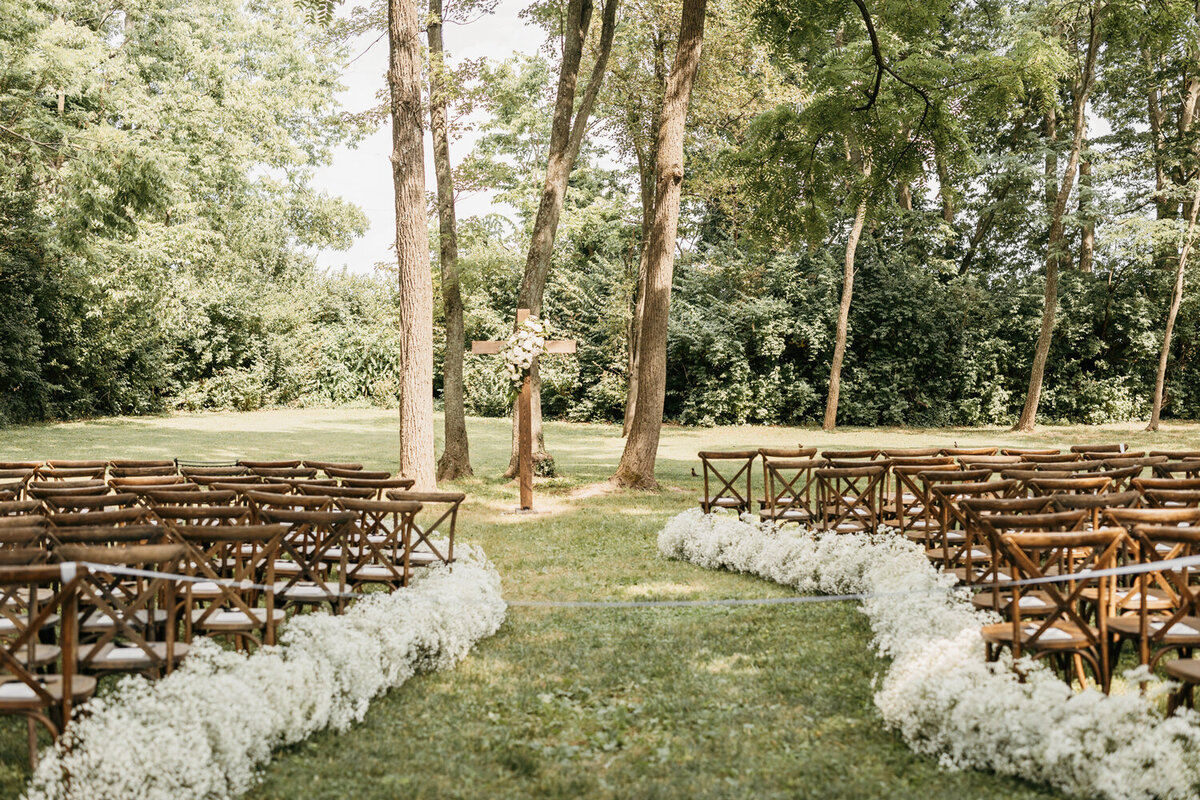 Elegant wedding ceremony setup at Ashford Acres Inn in Cynthiana, KY, featuring a wood cross adorned with white floral arrangements at the altar, framed by delicate baby's breath lining the aisle.