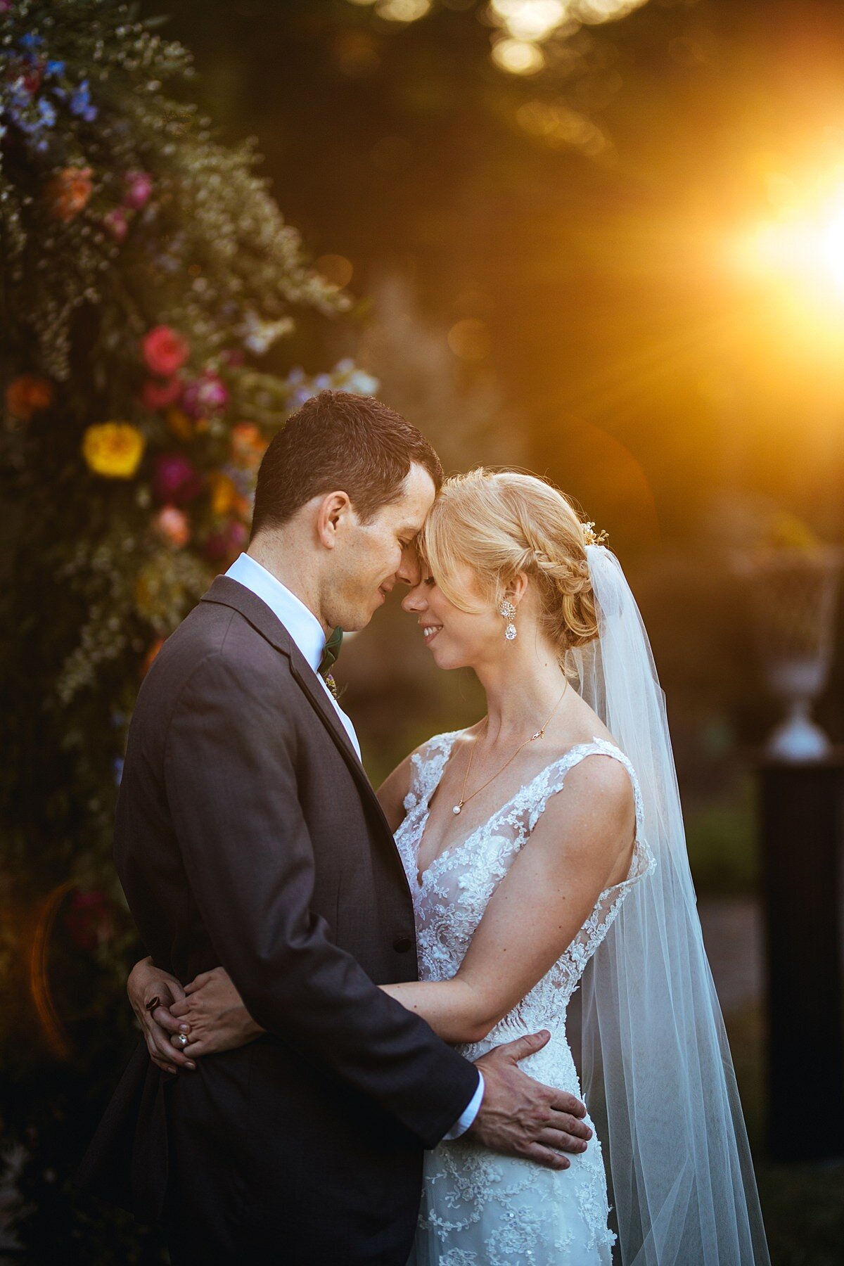 The bride, wearing a sleeveless lace wedding gown with a v-neckline and a long veil touching forheads with the groom wearing a charcoal gray suit as they embrace during golden hour as the sun sets at Cheekwood Botanical Garden