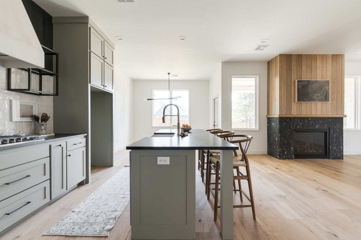 Open-concept kitchen and living space with sage green cabinetry, black countertops, and a modern gooseneck faucet island facing a wood-paneled fireplace with black marble surround and natural light from large windows.