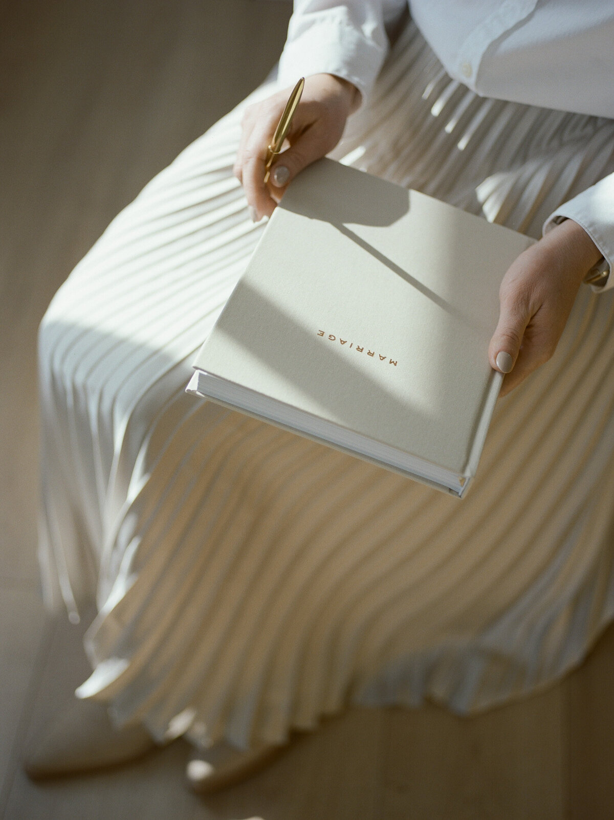 golden tones, directional light, woman in champagne pleated skirt holds beige linen journal with gold lettering, film product photography